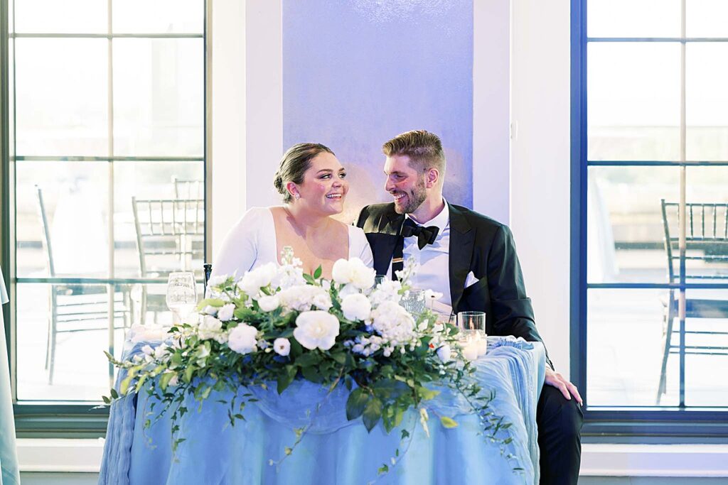 Couple smiles while sitting at table with blue tablecloth and white flowers