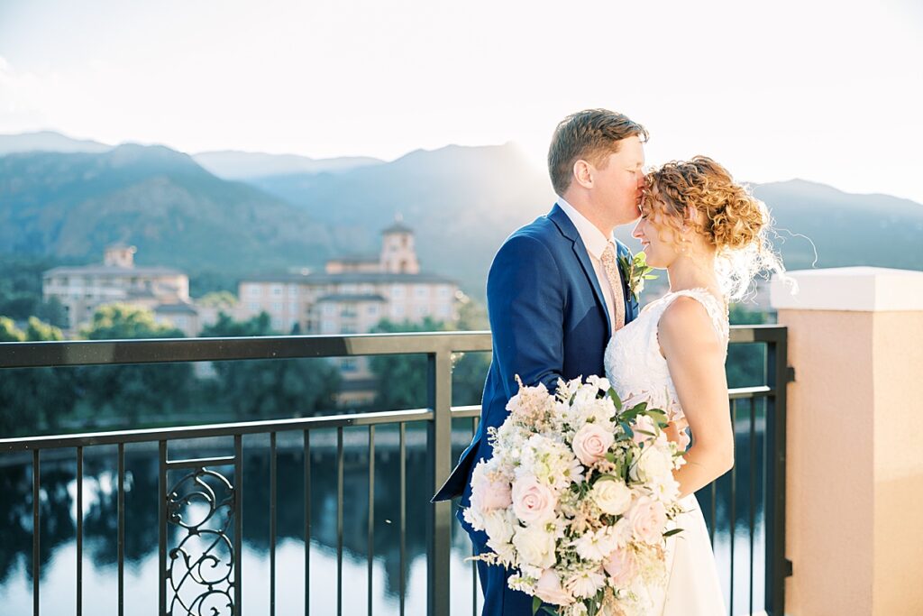 Groom in blue suit kissing bride on the forehead with mountains in background