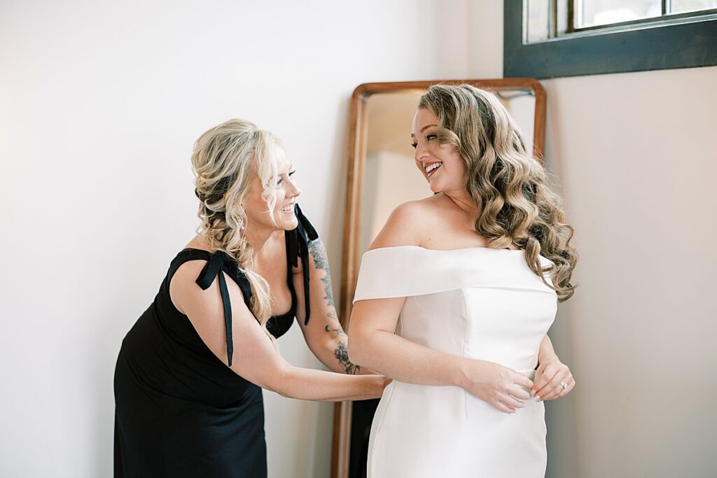 Bride smiles over her shoulder at bridesmaid zipping dress