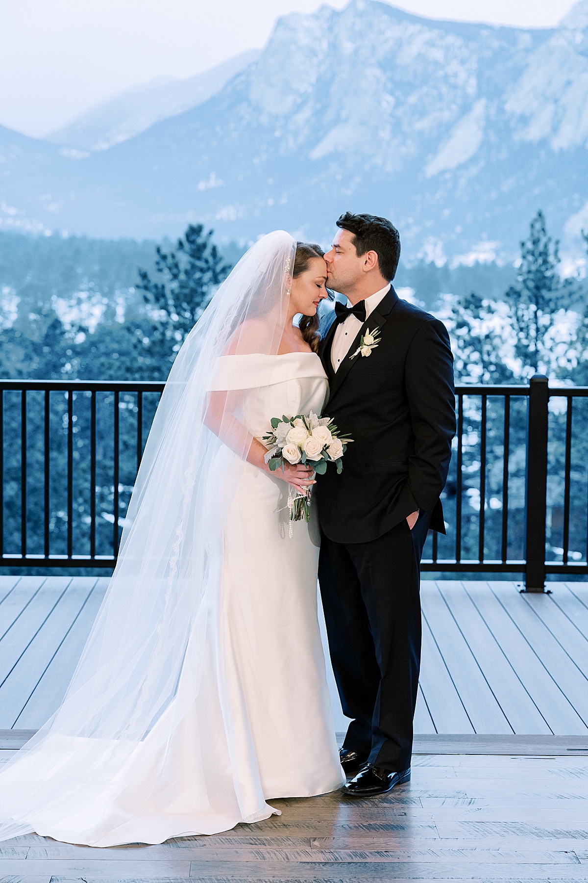 Groom kissing his bride on the forehead standing on deck with mountains