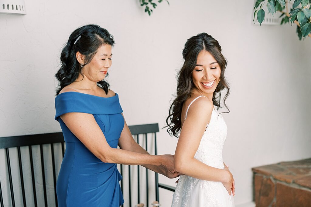 Bride looking over her shoulder at woman in blue dress zipping up her wedding dress