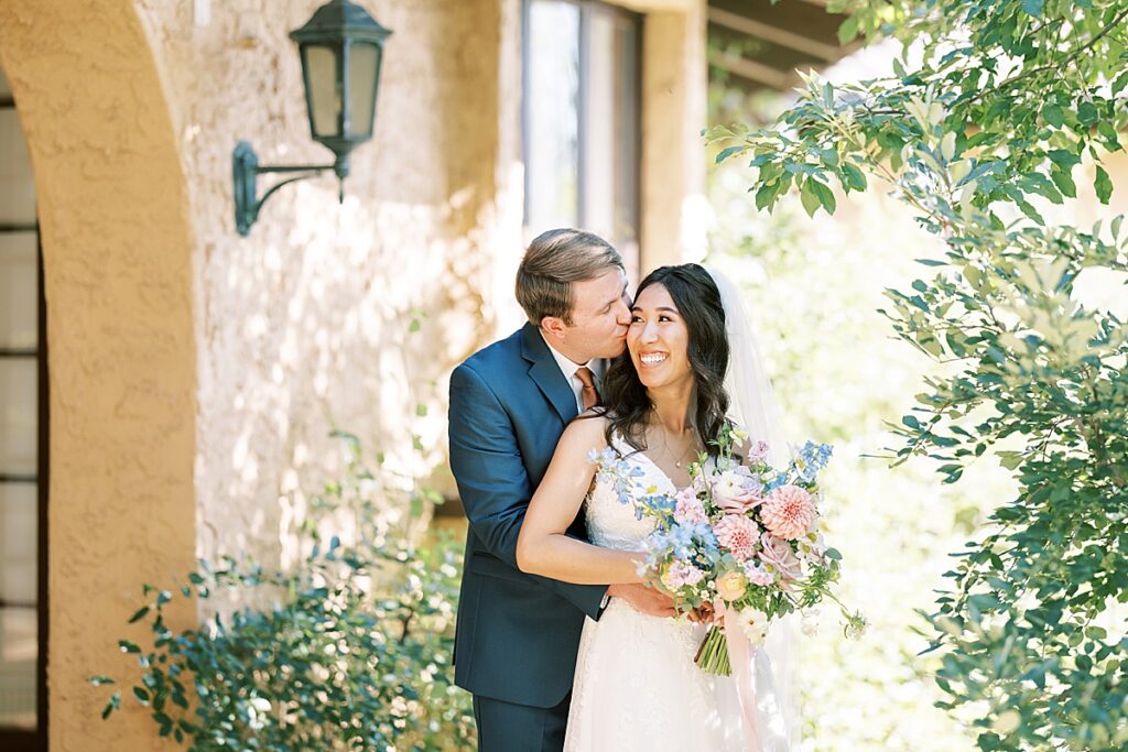 Groom in blue suit standing behind bride and kissing her on the cheek