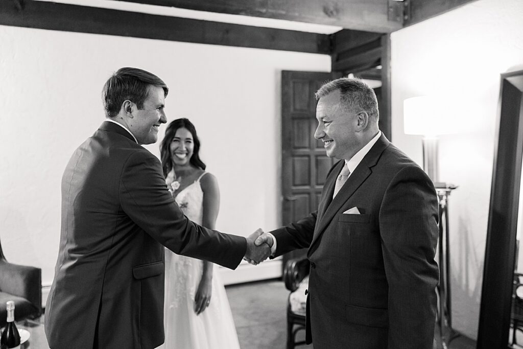 Black and white photo of two men shaking hands while bride smiles in background