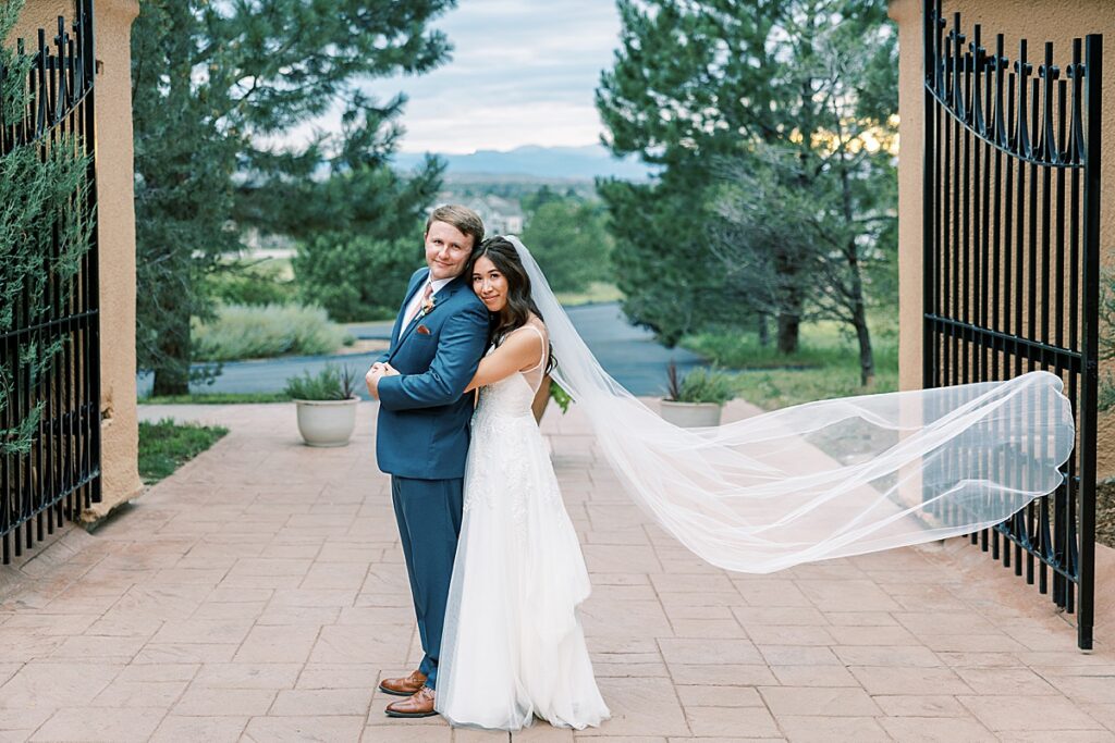 Bride hugs groom from behind with veil flying