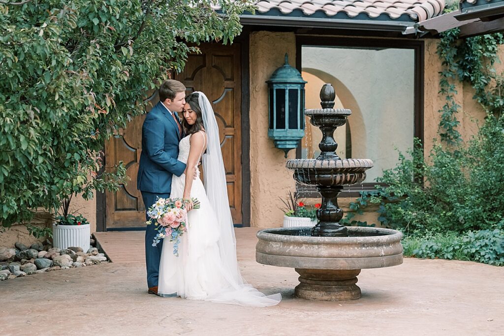 Bride looking down as groom kisses her on head in front of fountain