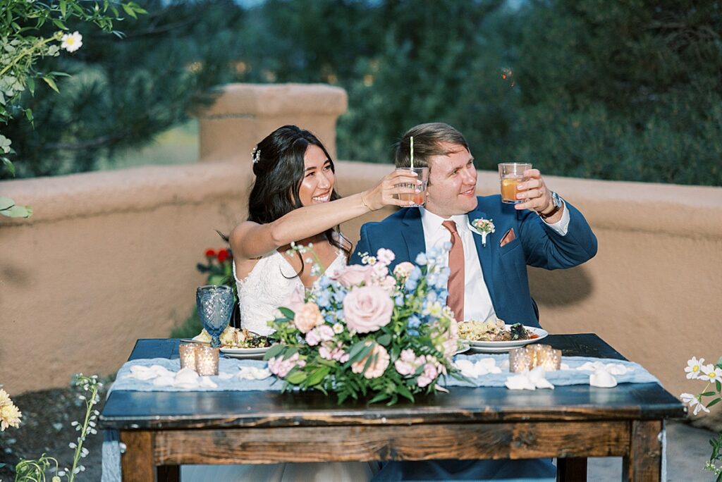 Bride and groom sitting behind table holding up glasses