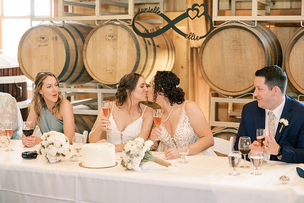 Two brides kissing while man in suit and woman in blue dress cheer at table