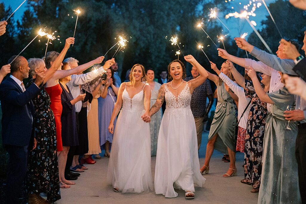 Two women in wedding dresses holding hands walking through sparklers