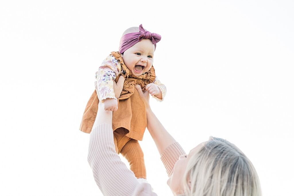 Woman in pink sweater holding a baby in orange jumpsuit and pink headband in the air